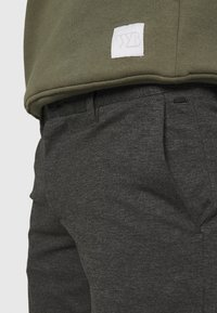 Olive green sweatshirt with a white logo patch, paired with dark gray textured pants featuring a side pocket and waistband detailing.