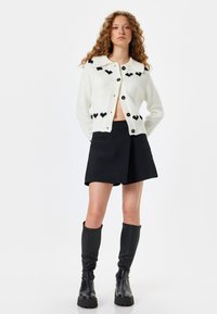Cárdigan blanco de punto con patrones de corazones negros, con cuello, cierre de botones, usado sobre una falda negra y botas negras hasta la rodilla.