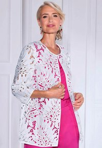 Weißer gehäkelter Cardigan mit Blatt-Ausschnittmustern. Lockere Passform, 3/4-Ärmel, über einem einfarbigen rosa Top getragen. Glatte Textur, leichtes Material.