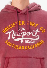 Sudadera burdeos con un bolsillo canguro, con bordados en amarillo y blanco: "Hollister Surf Co," "Newport Beach" y "Southern California."