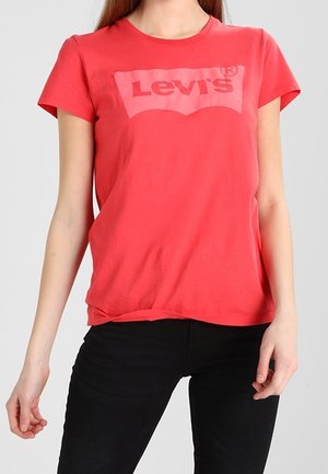 Kobieta w czerwonej koszulce z logo Levi's i czarnych spodniach, stojąca z luźno opuszczonymi ramionami na jednolitym jasnym tle.