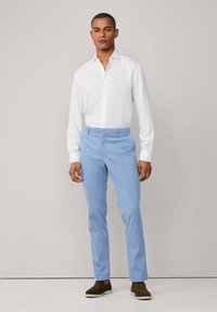 Man in een wit blouse met knopen, lichtblauwe broek en bruine instappers, staand tegen een effen lichtgrijze achtergrond.