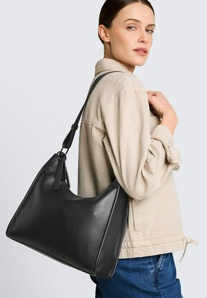 Frau trägt beige Jacke und blaue Jeans und hat eine große schwarze Leder-Schultertasche auf der rechten Schulter.