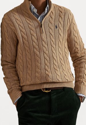 Homme portant un pull à col zippé en tricot torsadé de couleur beige sur une chemise bleu clair boutonnée, avec un pantalon foncé et une montre au poignet gauche.