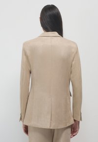 Blazer beige su misura con una texture liscia, rever a gola, due bottoni e una fessura sul retro per la mobilità, mostrato dal retro.