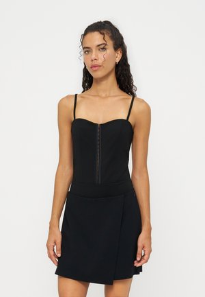 Jeune femme aux cheveux bouclés portant une robe noire sans manches avec de fines bretelles et une fermeture éclair devant sur fond blanc.