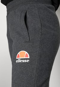 Dunkelgraue Fleece-Jogginghosen mit Seitentaschen, ausgestattet mit einem weißen und orangefarbenen "ellesse"-Logo am Bein. Sanfte Textur und lässiges Design.