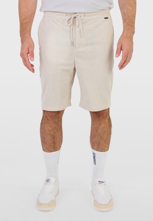 Hombre vistiendo pantalones cortos beige acanalados con cintura ajustable con cordón, calcetines blancos hasta el tobillo con texto azul y zapatillas blancas, de pie contra un fondo claro.