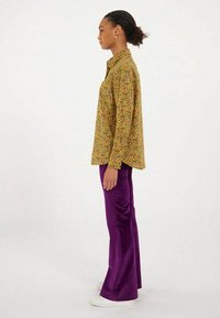 Chemise à manches longues jaune avec un imprimé floral multicolore, assortie à un pantalon évasé violet et des baskets blanches. Tissu lisse et léger.