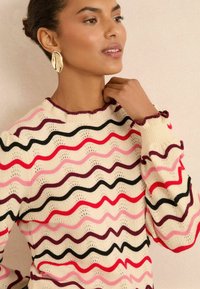 Eine Frau mit einem glatten Teint trägt einen wavy, mehrfarbigen Pullover und berührt ihren Hals. Sie hat große, modische Ohrringe und ein schwaches Lächeln.