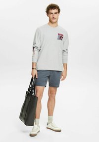 Lys grå langærmet skjorte med en brystlomme, der viser et "NY" grafisk design, parret med mørke shorts og hvide sneakers. Sort tote bag inkluderet.