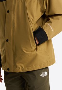Vandtæt jakke i sennepsgul med en tekstureret finish, med sorte accenter og logo. Inkluderer lommer og knaplukning.