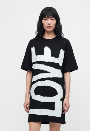 Femme aux longs cheveux noirs portant une robe t-shirt noire ample avec de grandes lettres abstraites blanches sur un fond uni.
