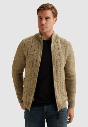 Homme portant un pull beige texturé zippé avec col et poignets côtelés, sur une chemise noire et un jean bleu, debout devant un fond uni.