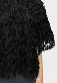 Μπλούζα με μαύρα κρόσσια και στρώσεις από υφασμάτινες λωρίδες με υφή. Το σχέδιο διαθέτει χαλαρή σιλουέτα και χαρακτηριστική κοπή στους ώμους, ενισχύοντας την κίνηση.