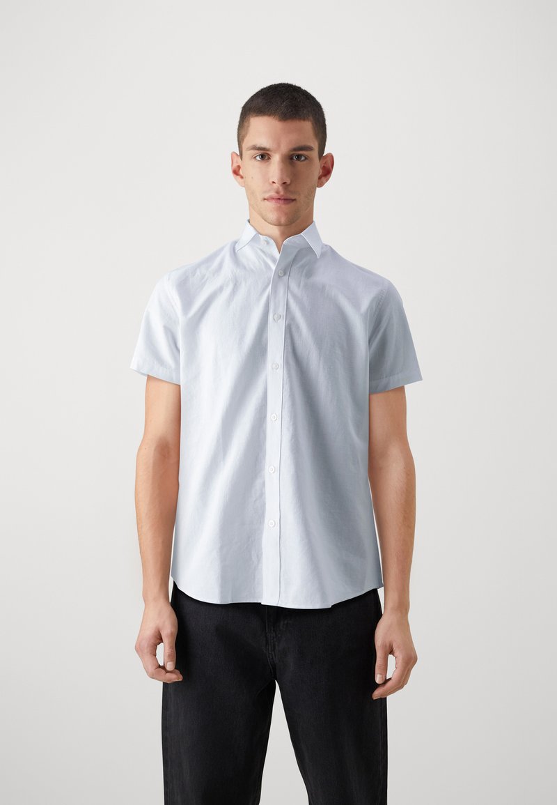 Camisa de manga corta azul claro con cuello abotonado, textura suave y cierre frontal de botones, combinada con unos jeans oscuros.