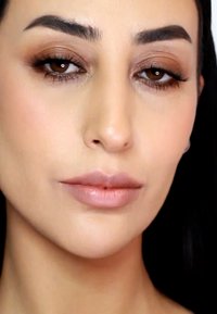 Primer plano de un rostro con piel suave y clara; ojos marrón claro, labios carnosos con un brillo nude. Las cejas están bien definidas y las pestañas son largas.