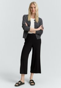 Cardigan en maille gris sur une chemise blanche, associé à un pantalon large noir. Des sandales noires avec un nœud mettent en valeur la tenue.