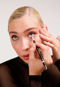 Makeupblyant i hånden, der påfører en lys nuance på det indre øje. Modellen har glat hud, blå øjne og lyse negle. Baggrunden er enkel og neutral.