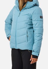 Femme portant une veste matelassée bleu clair à capuche avec des poches zippées et un pantalon noir, debout devant un fond uni.