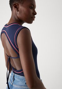 Merinavy bodysuit, jossa on korkea kaulus, punaisia yksityiskohtia ja avoin selkä. Varustettu solmittavalla sulkimella ja sileällä, joustavalla kankaalla. Yhdistetty vaaleisiin farkkushortseihin.