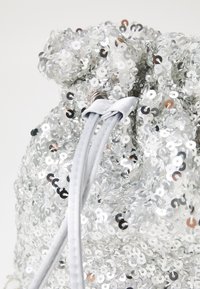 Hopea paljeteista valmistettu nyöripussukka, jossa sileä pinta. Siinä on pehmeä harmaa nauhakiinnitys ja pyöreä muoto.