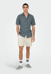 Camisa de botones de manga corta en gris oscuro, combinada con pantalones cortos beige. El modelo lleva zapatillas grises y calcetines de rayas blancas.