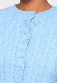 Gaiši zils adīts kardigāns ar kabeli rakstu un apaļu kaklu, aizverams ar apaļām gaiši zilām pogām.