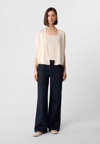 Cardigan beige chiaro, aperto davanti e con maniche lunghe, indossato sopra una maglietta aderente crema. Pantaloni in denim scuri a gamba larga, abbinati a ballerine color nude.