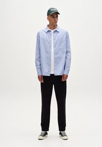 Chemise à rayures bleu clair et blanche avec un t-shirt blanc en dessous, pantalons noirs, baskets noir et blanc, casquette verte avec du texte.