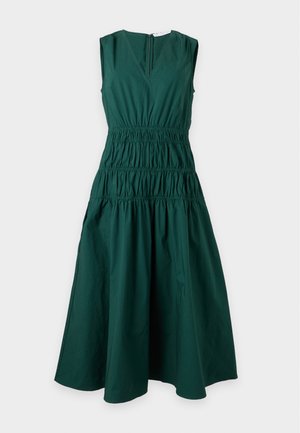 Woman wearing a sleeveless dark green midi dress with a V-neck and pockets, paired with brown flat sandals, standing against a white background.