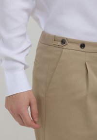 Beige Hosen mit strukturierter Oberfläche, ausgestattet mit einem doppelten Knopfverschluss und seitlichen Taschen, kombiniert mit einem weißen Langarmhemd.