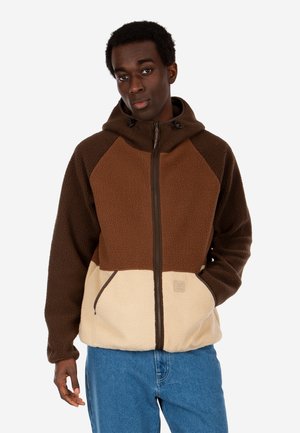 Man wearing a three-tone brown fleece jacket with a hood and blue jeans, standing with one hand in a pocket against a white background.