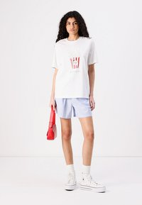 T-shirt bianco a maniche corte con grafica dei popcorn, abbinato a pantaloni corti a righe blu. Gli accessori includono una borsa rossa e sneakers bianche.