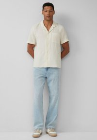 Korte mouwen crème button-up shirt met een zak, gecombineerd met lichtblauwe jeans en beige sneakers met witte accenten.