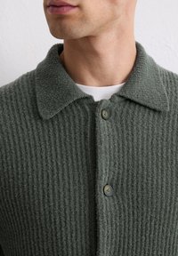 Groene gebreide cardigan met geribbelde textuur, ronde kraag en donkere knopen. Lichtgewicht materiaal, aansluitend ontwerp, geschikt voor layering.