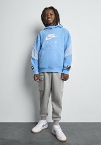 Jasnoniebieska bluza Nike z kieszenią z przodu i białym logo, zestawiona z szarymi spodniami dresowymi cargo oraz białymi sneakersami z niebieskimi akcentami.