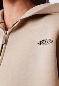 Sudadera con capucha beige con cremallera, confeccionada en tela suave, con un detalle de logo plateado en el pecho y capucha con cordón.