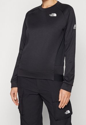 Persoon draagt een zwart The North Face sweatshirt met lange mouwen en zwarte cargobroek met het The North Face-logo op de zak, staand.