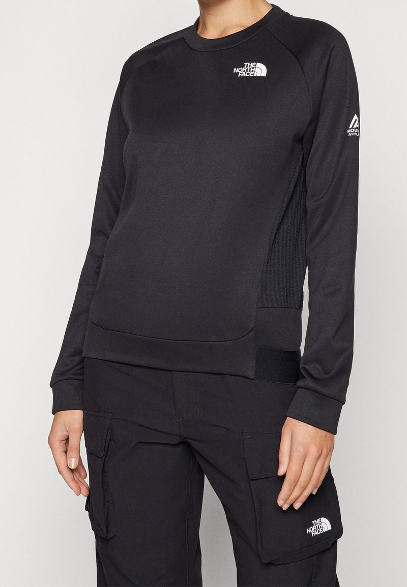 Personne portant un sweat-shirt à manches longues noir The North Face et un pantalon cargo noir avec le logo The North Face sur la poche, debout.