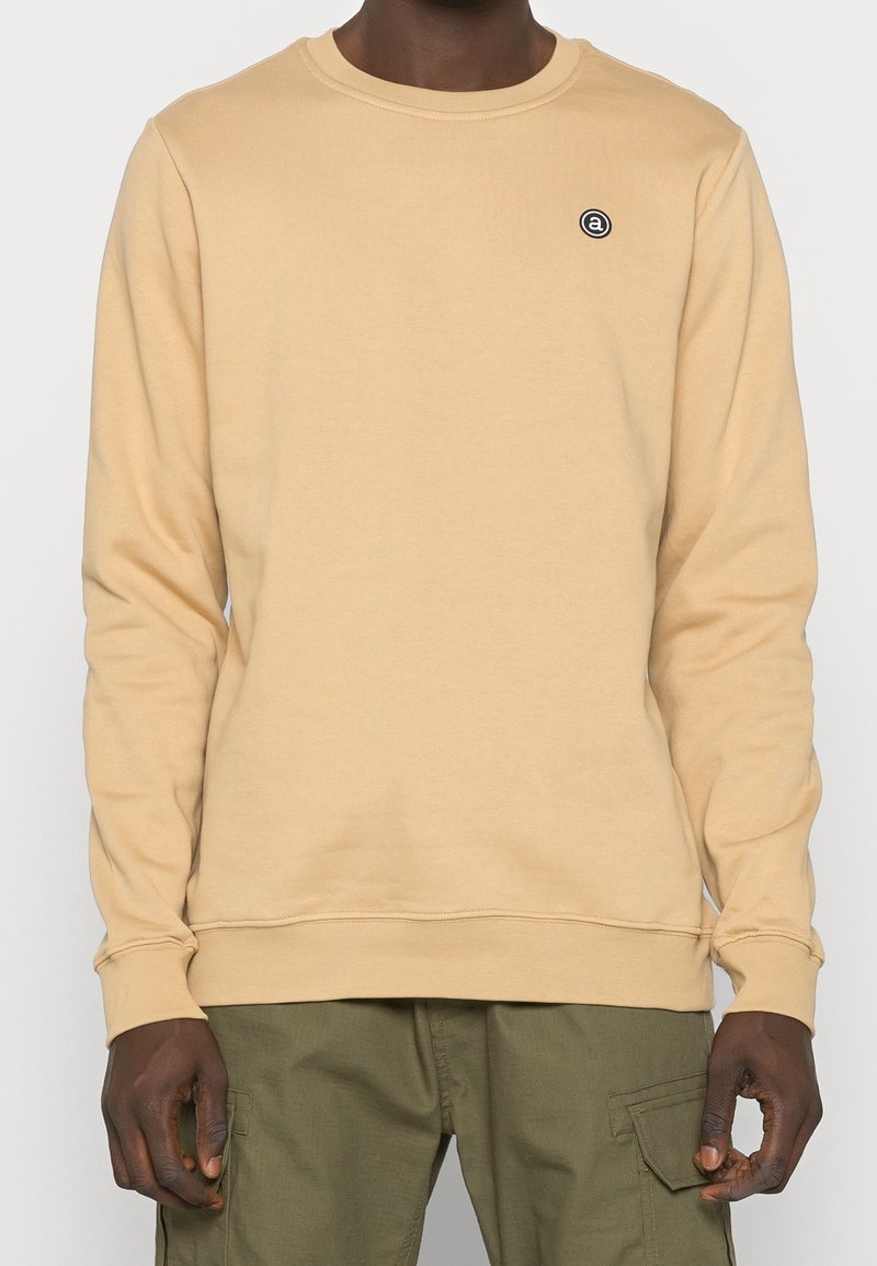 Sudadera beige claro con cuello redondo, mangas largas, puños y dobladillo acanalados. Presenta un pequeño logo circular negro en el lado izquierdo del pecho.