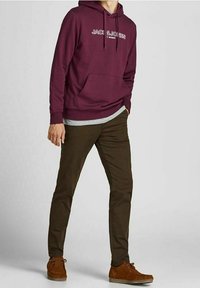 Sudadera con capucha color burdeos con bolsillo frontal y logo blanco, combinada con pantalones chinos marrón oscuro y zapatos de ante marrones. El modelo posa sobre un fondo neutro.