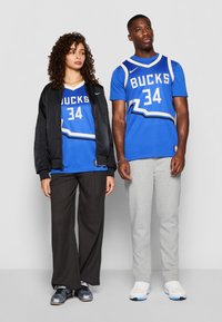 Mėlyni krepšinio marškiniai su užrašu "BUCKS" ir numeriu "34", derinami su juoda bomber striuke ir pilkomis treniruočių kelnėmis, vilkimo su sportiniais batais.
