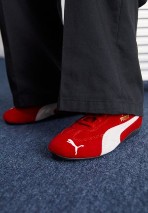Zapatillas de ante rojas con acentos blancos, que cuentan con material texturizado, punta redonda y un logo dorado en el lateral, combinadas con pantalones negros.