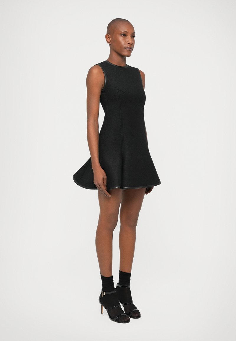 Robe noire sans manches avec jupe évasée, fabriquée à partir d'un tissu texturé, présentant un corsage ajusté et un ourlet contrasté. Portée avec des bottines à talons noires.