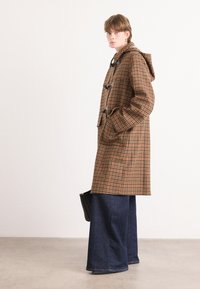 Bruin geruite jas met een capuchon, grote voorzakken en zwarte togglesluitingen, gedragen met wijduitlopende donkerdenim jeans.