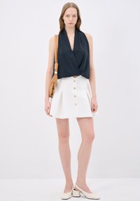 Top senza maniche navy con design drappeggiato, gonna bianca con bottoni davanti e pieghe, abbinata a scarpe slingback crema e borsa a tracolla beige.