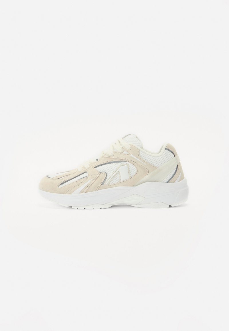 Beige en witte sportschoen van suede en mesh materialen, met een ronde neus, veters en textuuraccenten aan de zijkanten.