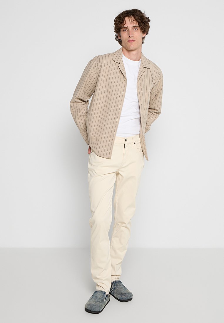 Lichtbeige gestreepte blouse over een wit T-shirt, gecombineerd met crèmekleurige broeken en grijze instappers. Eenvoudig ontwerp, casual pasvorm.