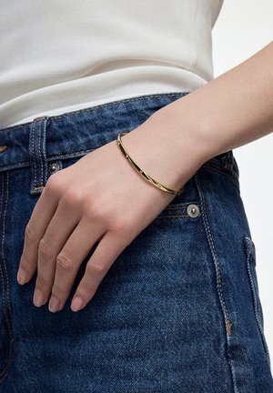 Złota metalowa bransoletka bangle o gładkiej powierzchni i skręconym wzorze. Noszona na ręku odpoczywającym na dżinsowych spodniach.